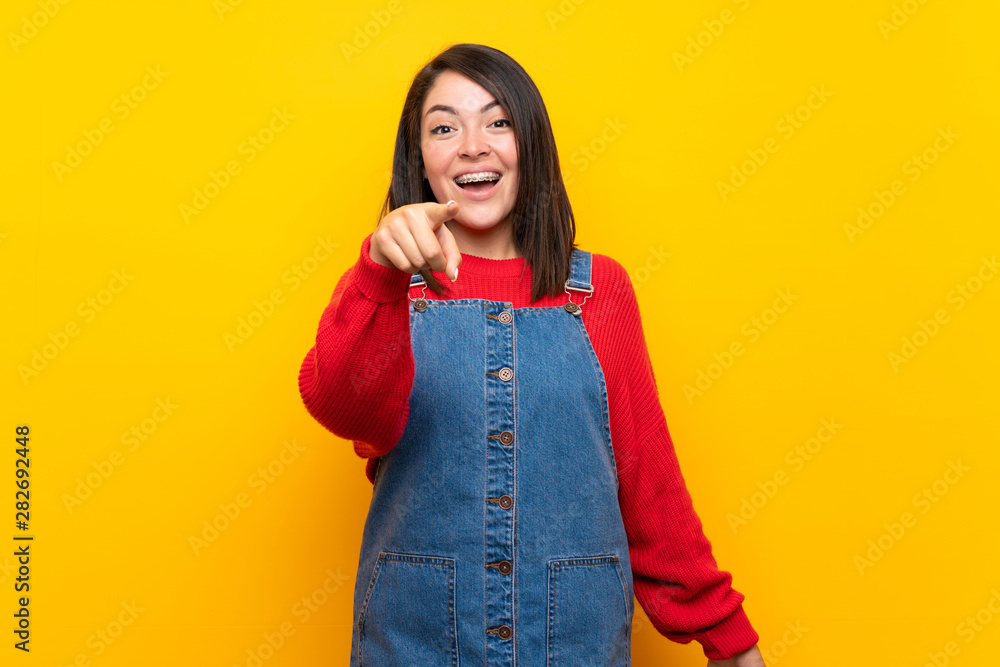Young Mexican woman with overalls over yellow wall surprised and pointing front Young Mexican woman with overalls over yellow wall surprised and pointing front