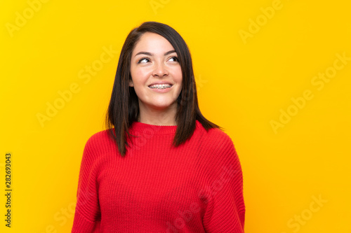 Young Mexican woman with red sweater over yellow wall laughing and looking up