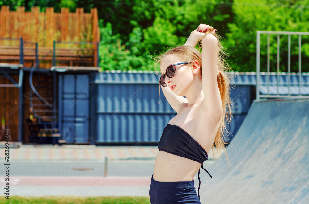 Slender teenage girl in a sports top and shorts on a playground for skaters in sunglasses