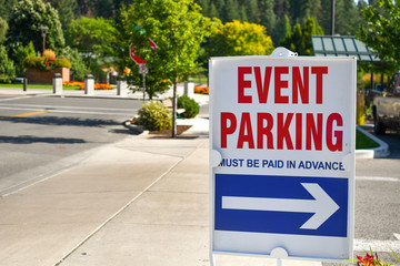 A red, white and blue parking sign on a street sidewalk points towards event parking on a sunny day in a non-descript mountain town.