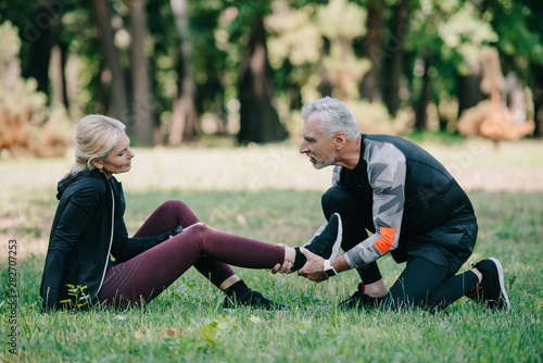 attentive mature sportsman touching injured leg of sportswoman sitting on lawn