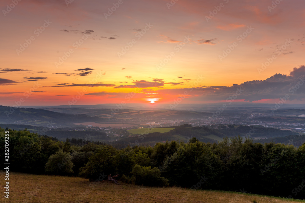 Sunset with view on landscape with fully colored clouds and orange sun goes down and city Valasske Mezirici captured during summer late time