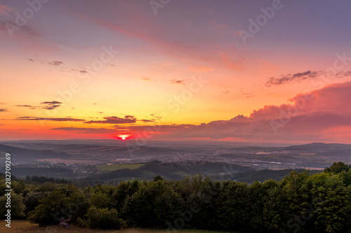Sunset with view on landscape with fully colored clouds and orange sun goes down and city Valasske Mezirici captured during summer late time