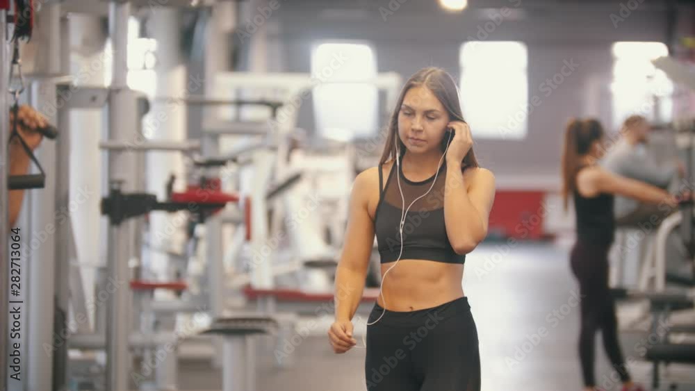 An athlete woman walking in the gym and putting on a headphones - get her hands in the handles and start training An athlete woman walking in the gym and putting on a headphones - get her hands in the handles and start training