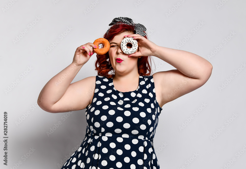 Portrait of beautiful cheerful fat plus size woman pin-up wearing a polka-dot dress isolated over light background, eating a donut Portrait of beautiful cheerful fat plus size woman pin-up wearing a polka-dot dress isolated over light background, eating a donut