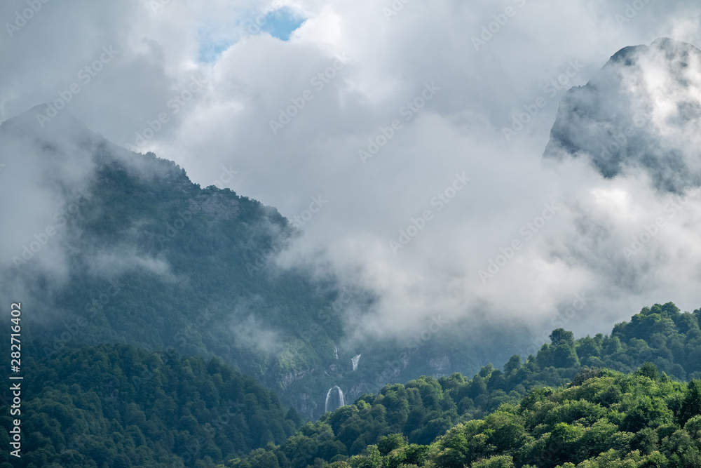 Green mountains with a high waterfall in fog and clouds.