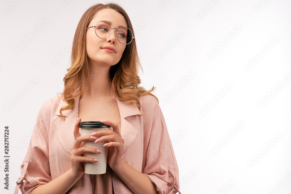 Young attractive woman with dark blond curly hair wearing stylish round glasses and fashionable pink trench coat holds coffee mug, looks aside with slight smile and pleasant dreamy expression. Young attractive woman with dark blond curly hair wearing stylish round glasses and fashionable pink trench coat holds coffee mug, looks aside with slight smile and pleasant dreamy expression.