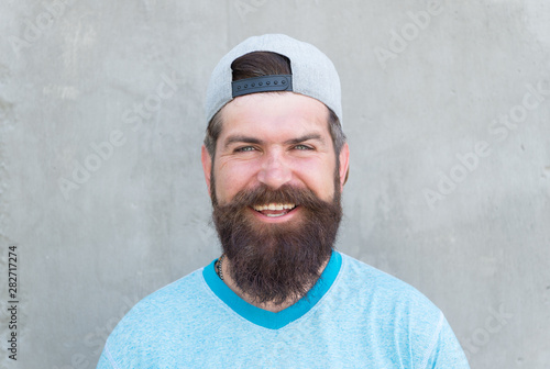 Keeping hair healthy. Happy hipster with shaped beard and styled hair wearing baseball cap. Bearded man smiling with unshaven face hair. Hairy guy with stylish beard and mustache hair on grey wall