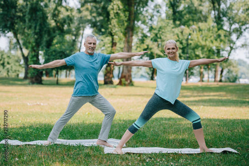 positive mature man and woman practicing warrior poses in park on lawn