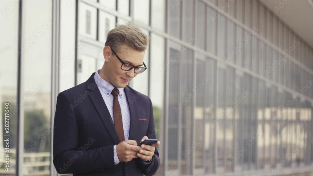 Happy businessman smiling as he reads a text message standing outside the office. Male executive manager in trendy clothes using smartphone outdoors. Modern technology concept Happy businessman smiling as he reads a text message standing outside the office. Male executive manager in trendy clothes using smartphone outdoors. Modern technology concept