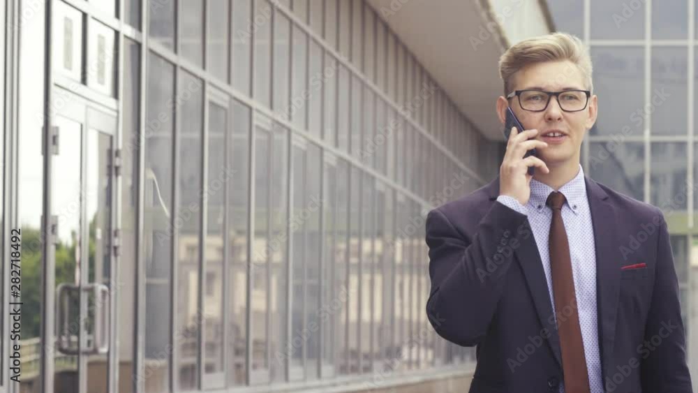 Happy attractive caucasian businessman walking outside near office building and talking on mobile phone. He comes in and out of focus passing by camera. Happy attractive caucasian businessman walking outside near office building and talking on mobile phone. He comes in and out of focus passing by camera.