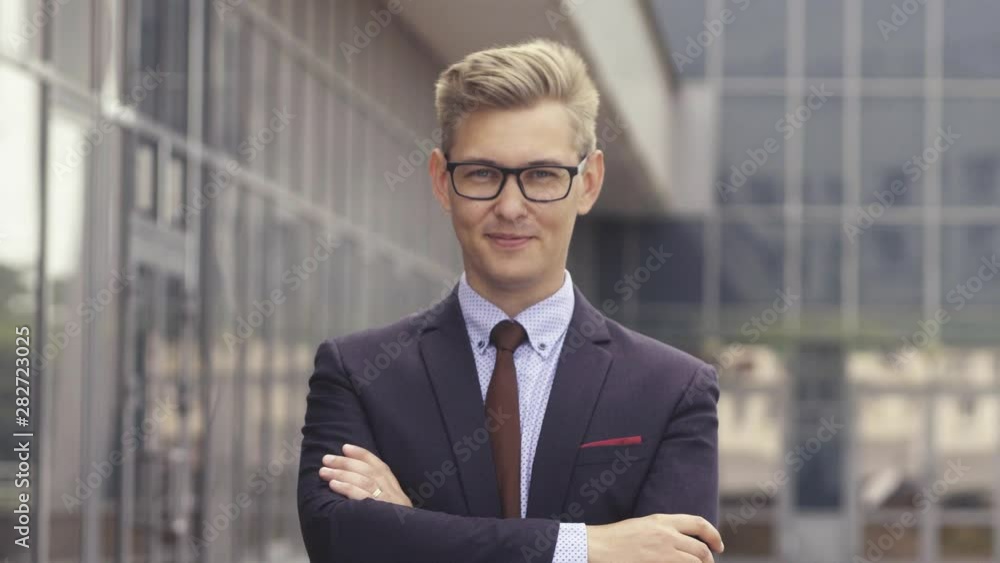 Portrait man in suit in front of office building. Handsome businessman outdoor. Male business person stands on street and looking away. Businessman dressed in formal clothes and glasses. Portrait man in suit in front of office building. Handsome businessman outdoor. Male business person stands on street and looking away. Businessman dressed in formal clothes and glasses.