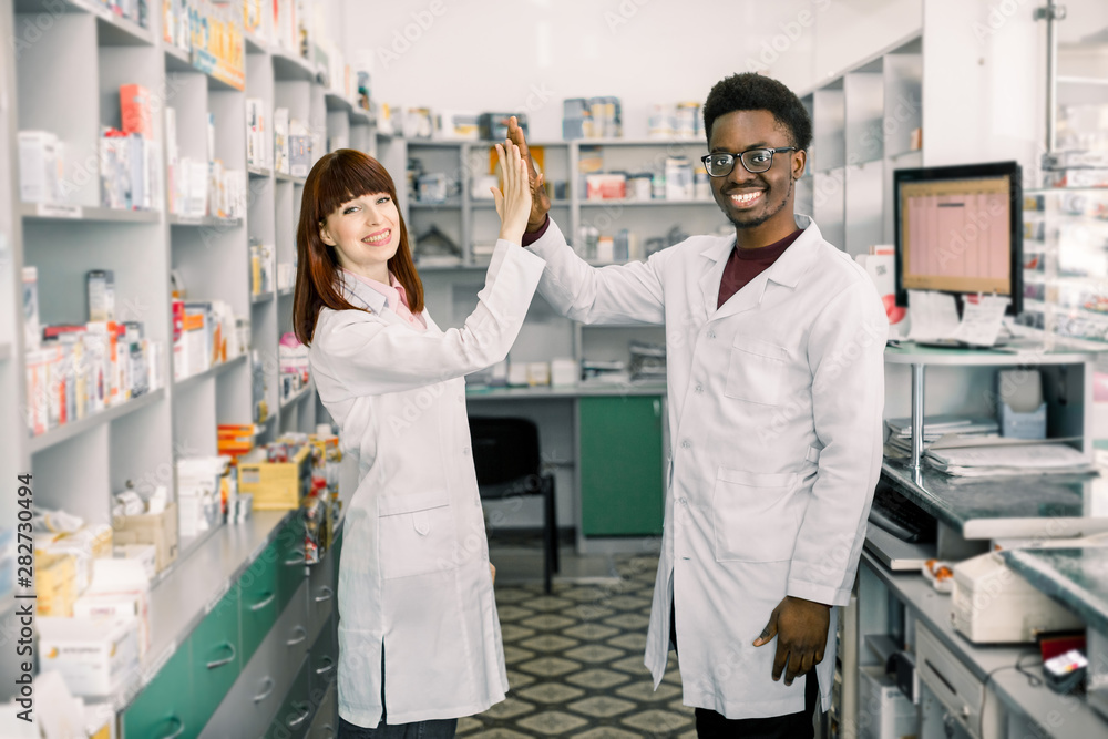 Obraz premium Female and male pharmacists in pharmacy. African man and Caucasian woman giving five, working at modern pharmacy