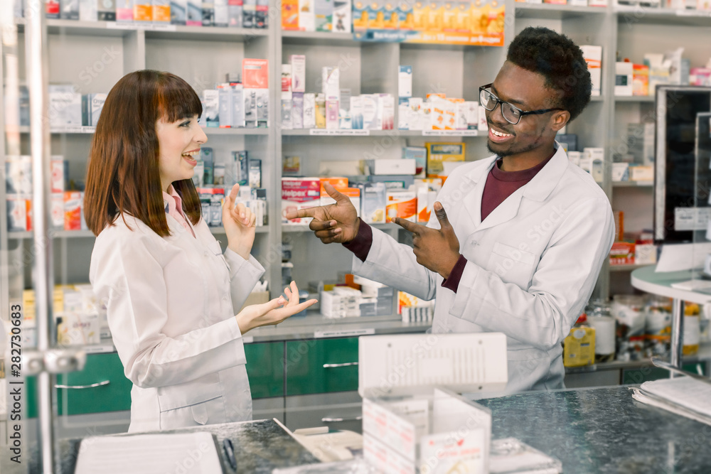 Portrait of multiethnical male and female pharmacists standing near pharmacy counter, talking, laughing and gesturing each other