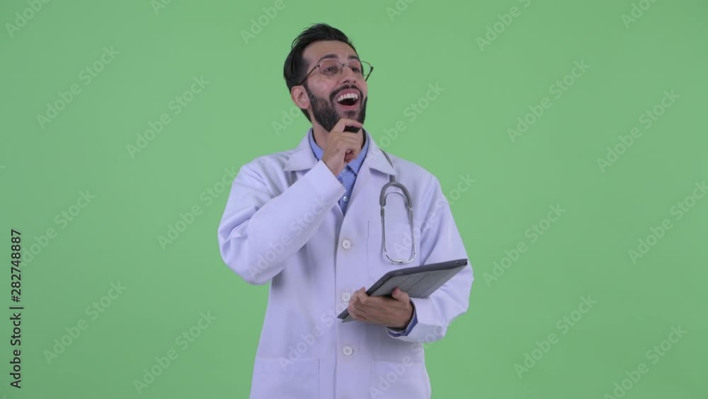 Happy young bearded Persian man doctor thinking while using digital tablet Happy young bearded Persian man doctor thinking while using digital tablet