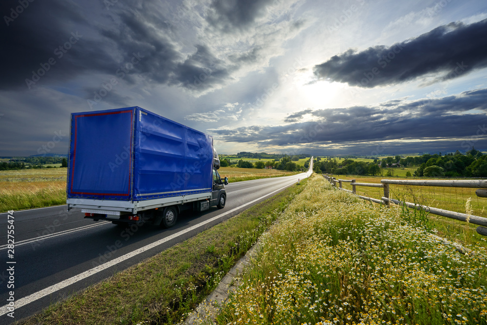 Delivery van driving on the asphalt road in rural landscape at sunset with dramatic clouds