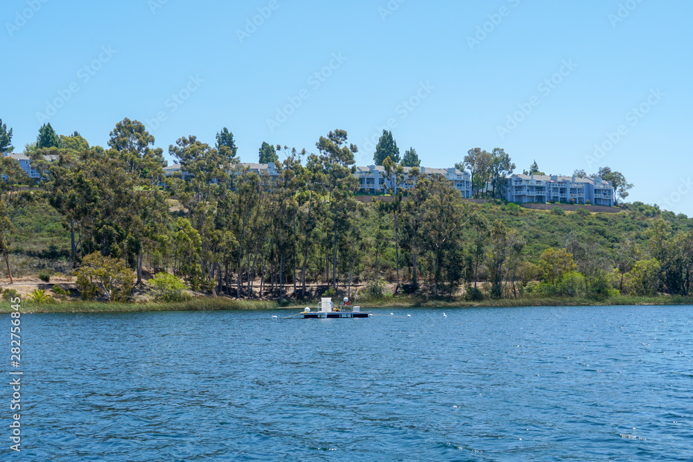 Big lake with blue water, trees and native wetland plants and villa on the cliff with blue sky. Miramar reservoir in the Scripps Miramar Ranch community, San Diego, California
