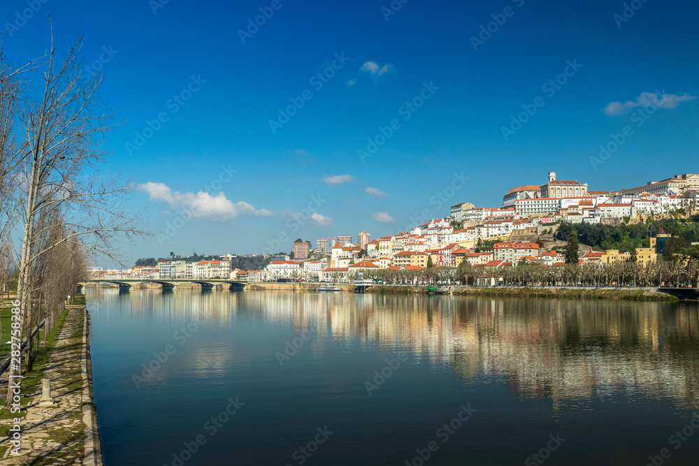 Cityscape of Coimbra in Portugal with the Mondego river in the foreground and the reflection of the city in its waters on a sunny winter morning.