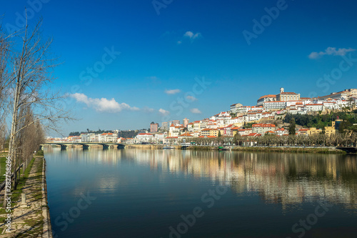 Cityscape of Coimbra in Portugal with the Mondego river in the foreground and the reflection of the city in its waters on a sunny winter morning.