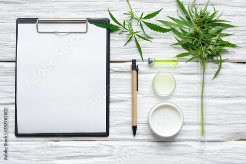 Cannabis cosmetic cream in a jar and a blank paper page with a copy space and a green hemp leaf on a white wooden table background. © Natali