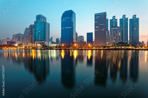 Night skyline of modern lakeside skyscrapers with glass curtain walls and dazzling city lights reflected in the smooth lake water in beautiful Benjakiti Park at blue dusk, in Bangkok, Thailand, Asia