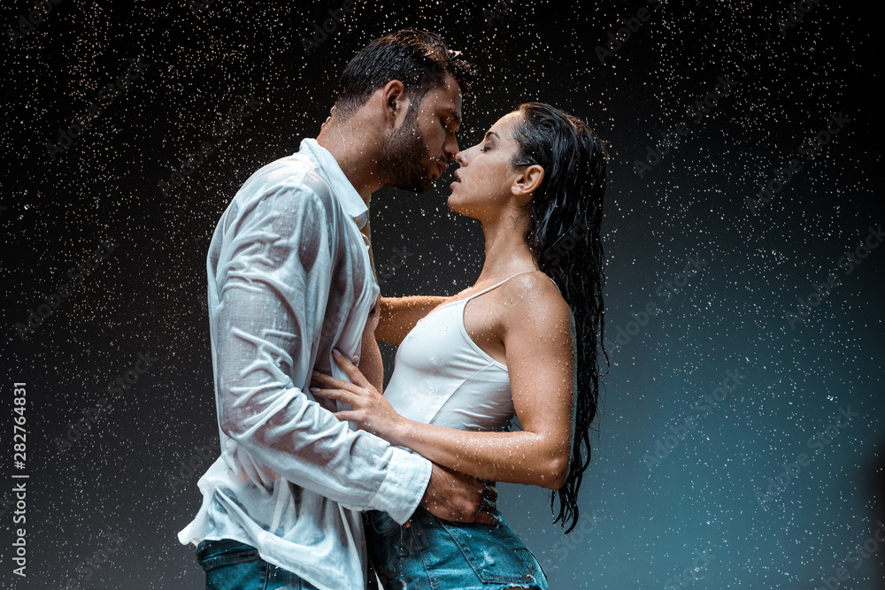 side view of handsome man kissing sexy wet girlfriend under raindrops on black - obrazy, fototapety, plakaty 