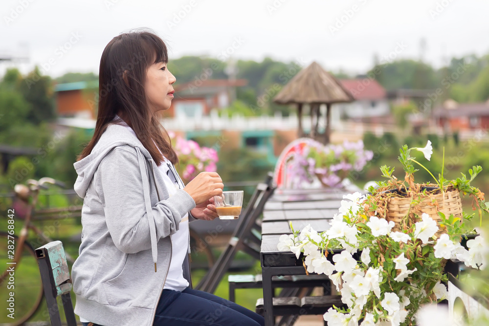 Asian woman is sitting on a wooden table waiting to watch the sunrise in the morning at rural , Thailand. The elderly Asian people are drinking hot coffee on a wooden table and watching the sunrise.