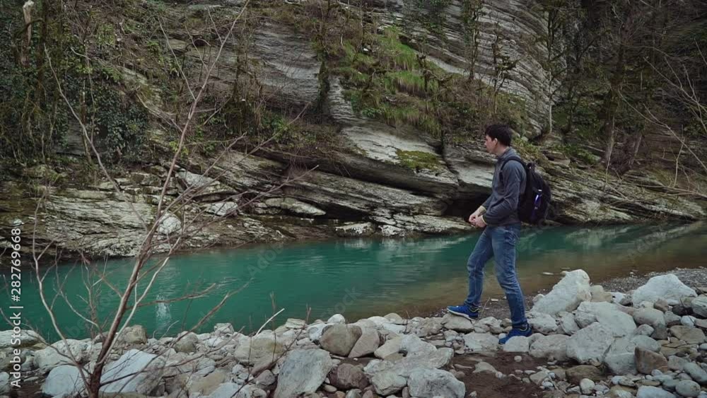 Tall handsome guy with a backpack stands on the bank of a clean mountain river and throws stones into the water.