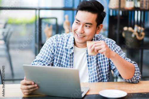 Asian man using laptop and drinking coffee in coffee shop cafe working online freelance business