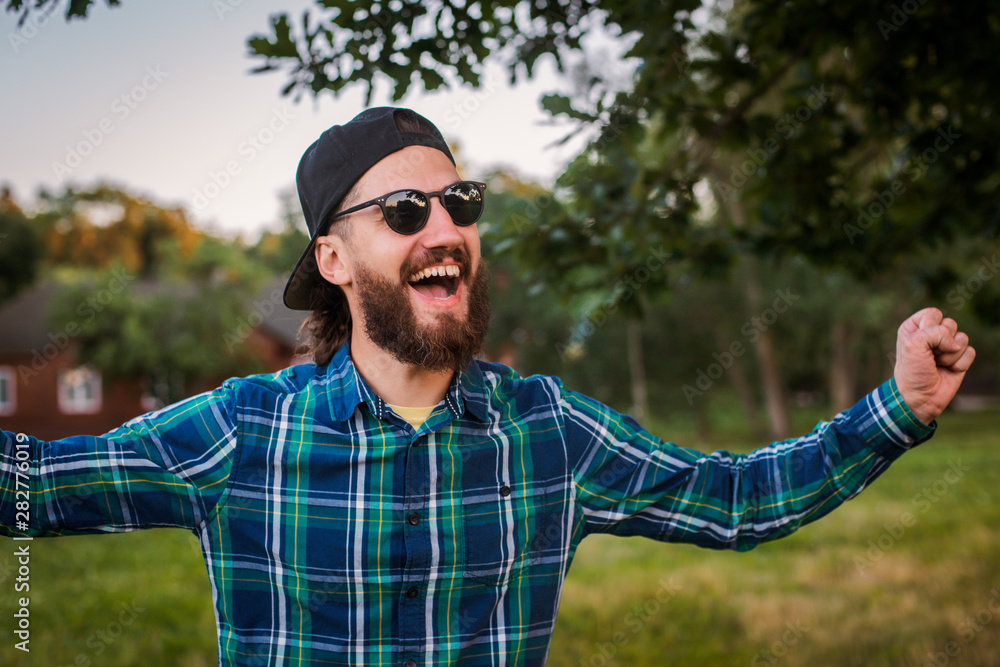 Obraz premium Portrait of a young hipster man in sunglasses and baseball cap posing in the backyard outdoors.
