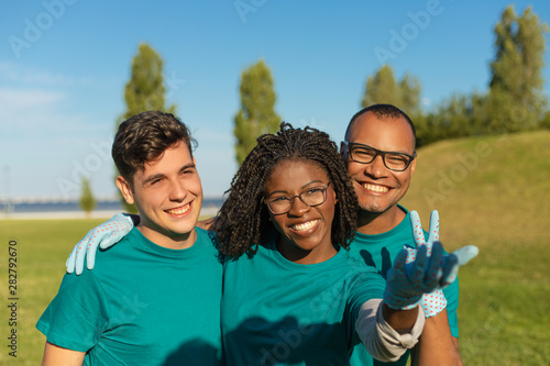 Happy volunteers rejoicing at something outside. Young men and woman in uniforms standing on grass, hugging, looking and pointing away, smiling and laughing. Volunteer team concept