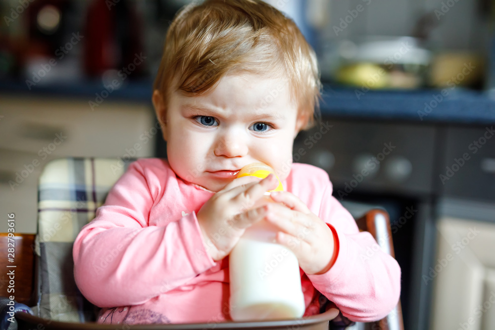 Cute adorable baby girl holding nursing bottle and drinking formula milk. First food for babies. New born child, sitting in chair of domestic kitchen. Healthy babies and bottle-feeding concept Cute adorable baby girl holding nursing bottle and drinking formula milk. First food for babies. New born child, sitting in chair of domestic kitchen. Healthy babies and bottle-feeding concept