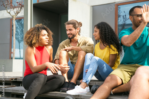 Excited black girl telling funny story to her friends. Multiethnic group of young people sitting outside, talking, chatting, laughing. Friendship concept