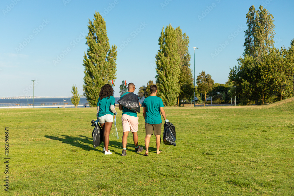 Outdoor cleaners removing trash from city lawn. Back view of woman and men walking through, holding rakes and plastic bags. Outdoor rubbish collection concept