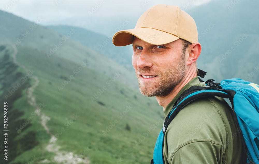 Portrait of backpacker man in baseball cap walking by the foggy cloudy weather mountain range path with backpack. Active sports backpacking healthy lifestyle concept.