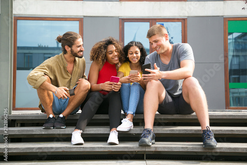 Happy joyful friends sitting on staircase and holding mobile phones. Interracial girls and guys showing smartphone screens to each other. Communication concept