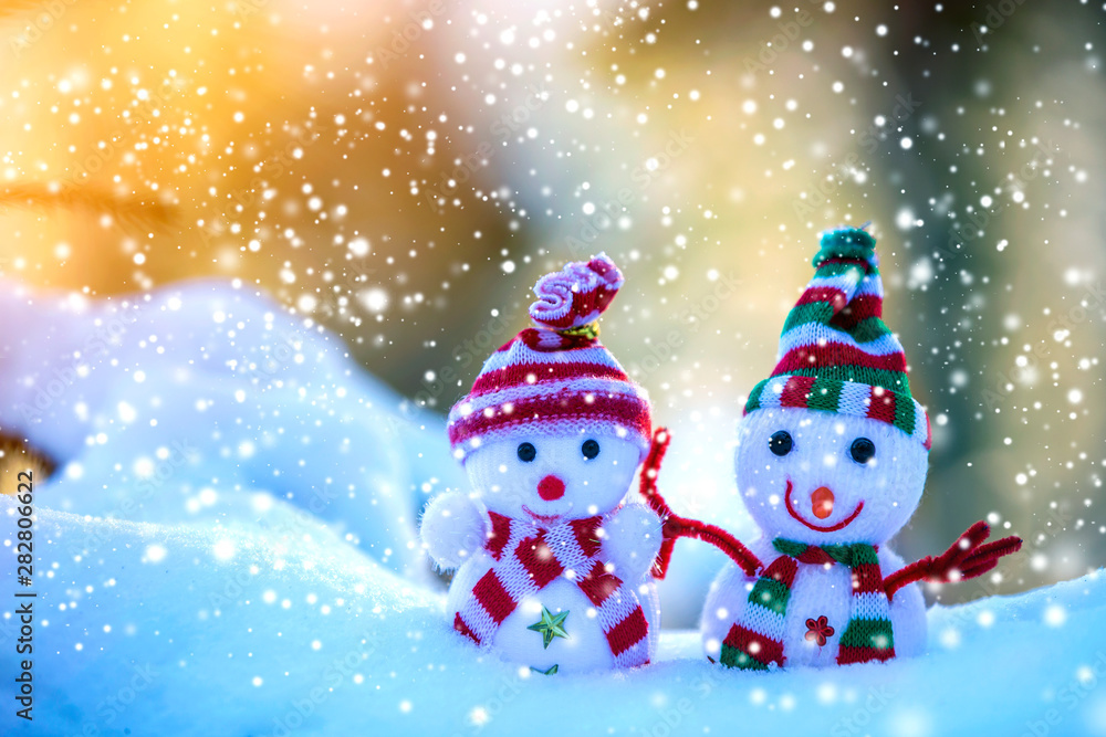 Two small funny toys baby snowman in knitted hats and scarves in deep snow outdoors on bright blue and white copy space background. Happy New Year and Merry Christmas greeting card.