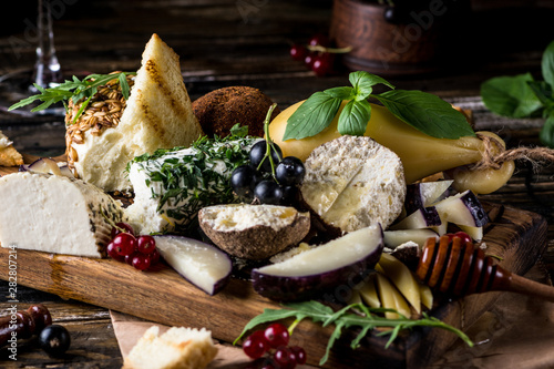 Cheese appetizer selection. Red currant, honey, basil, grapes and nuts on rustic wooden board over wooden concrete backdrop, top view. Belper Knolle, goat cheese, Scamorza. Cheese board, cheese plate.