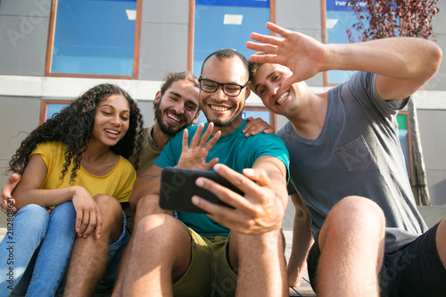 Happy friends talking to another friend through video call on mobile phone. Young men and woman sitting outside, gesturing, laughing, smiling at smartphone screen. Video chat concept