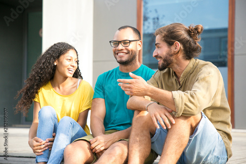 Happy multiethnic friends chatting outside. Cheerful mix raced young men and woman sitting outdoors, laughing, talking, discussing gossips. Leisure and company concept