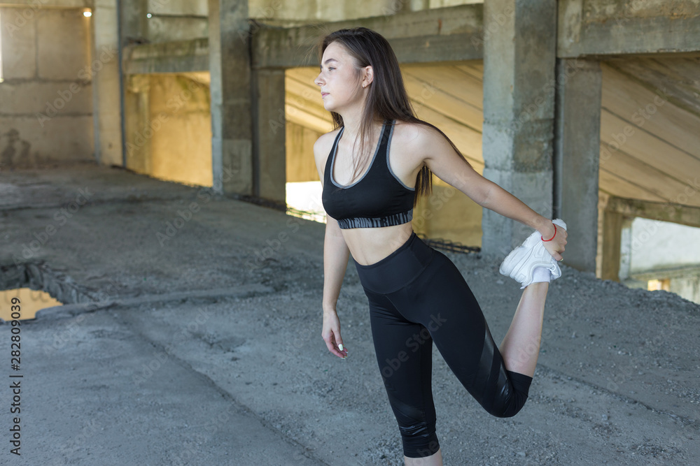 Slim athletic girl performs stretching exercises on the roof of an unfinished building, urban background.