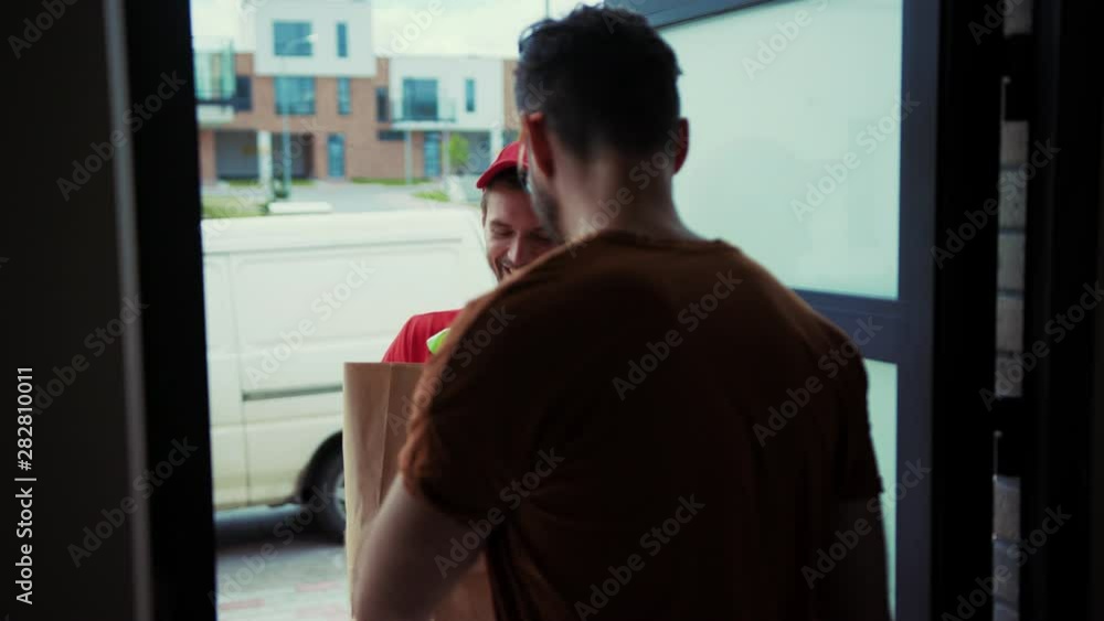 Attractive man opening the door to courier receiving delicious fresh food from supermarket. Close-up guy paying with cash taking order from smiling happy delivery man. Food home delivery. Attractive man opening the door to courier receiving delicious fresh food from supermarket. Close-up guy paying with cash taking order from smiling happy delivery man. Food home delivery.