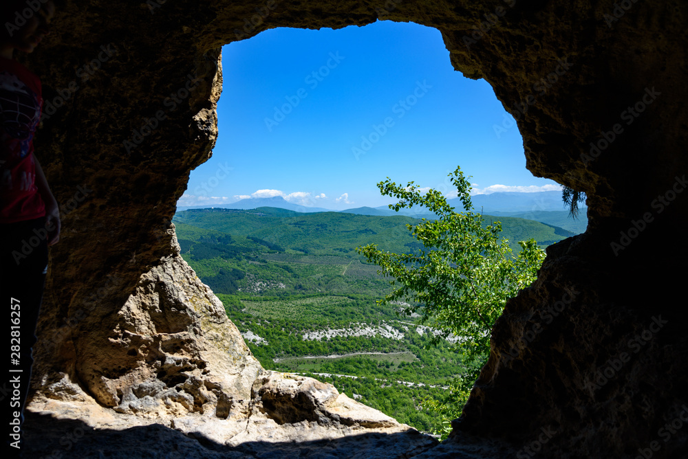 View of the mountain covered with a green carpet of trees and grass, through a window from a cave, on a bright sunny day, with clouds in the sky. Spring view of the Crimean mountains.