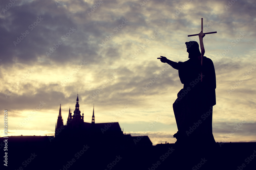 Naklejka premium Silhouette of St. John the Baptist statue on Charles Bridge in Prague with St. Vitus Cathedral in background at sunset.