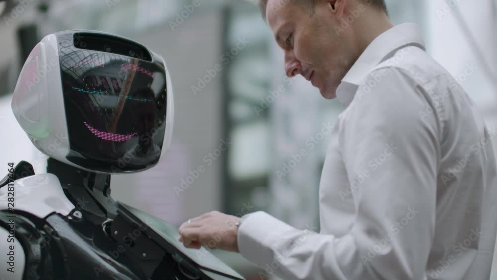 A man stands with a robot bot and asks him questions and asks for help by clicking on the screen on the robot body. A man stands with a robot bot and asks him questions and asks for help by clicking on the screen on the robot body.