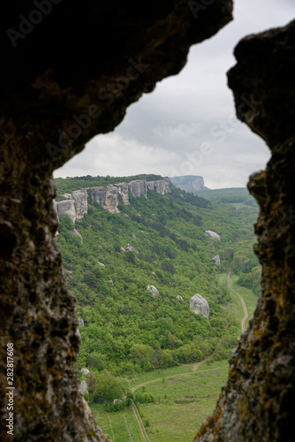 View of the mountain covered with a green carpet of trees and grass, through a window from a cave, on a bright sunny day, with clouds in the sky. Spring view of the Crimean mountains.