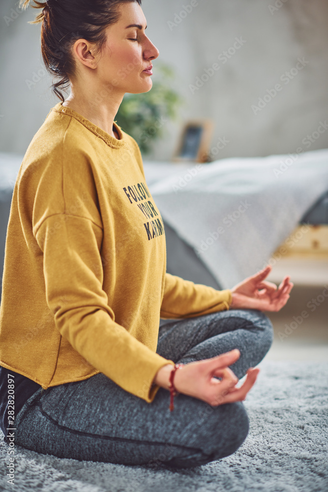 Beautiful Caucasian brunette sitting on rug in bedroom in Lotus yoga posture.