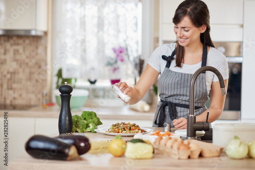 Cute Caucasian brunette in apron standing in kitchen and adding salt in fresh delicious pasta. Preparation of italian food concept.