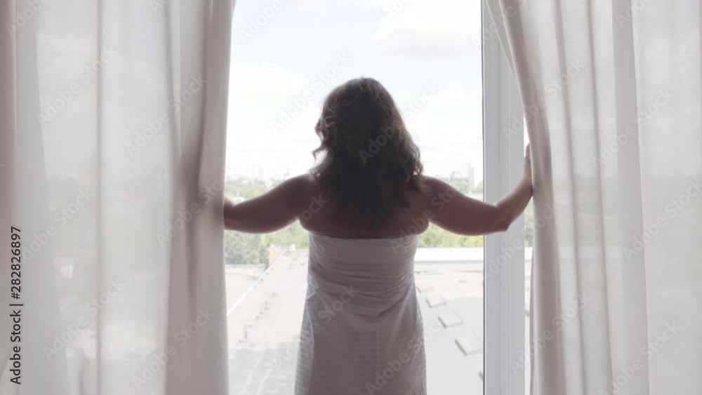 Rear back view at rich mature woman opening curtain lace standing in luxury apartment home or modern hotel looking through window enjoying wellbeing. Happy caucasian woman greets new day. Rear back view at rich mature woman opening curtain lace standing in luxury apartment home or modern hotel looking through window enjoying wellbeing. Happy caucasian woman greets new day.