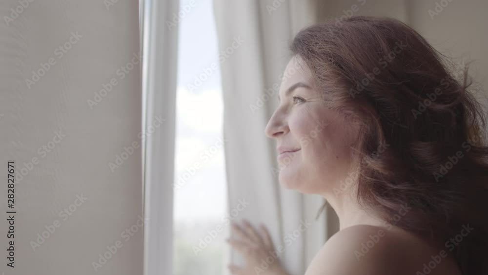 Beautiful mature woman opening curtain lace standing in luxury apartment home or modern hotel looking through window enjoying wellbeing. Happy caucasian woman greets new day. Beautiful mature woman opening curtain lace standing in luxury apartment home or modern hotel looking through window enjoying wellbeing. Happy caucasian woman greets new day.